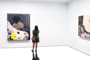 A woman with brunette, shoulder-length hair, wearing a grey blouse and skirt and leather handbag is seen in a bright, bare gallery setting save for two large, colorful paintings hanging on the walls, one of which she is seen from behind as she views it