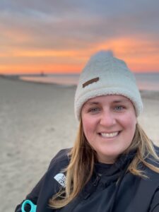 A woman with shoulder length, sandy blonde hair, a fair complexion and wearing a dark-colored North Face windbreaker and white, wool winter cap is standing on a beach at sunset looking to the camera and smiling