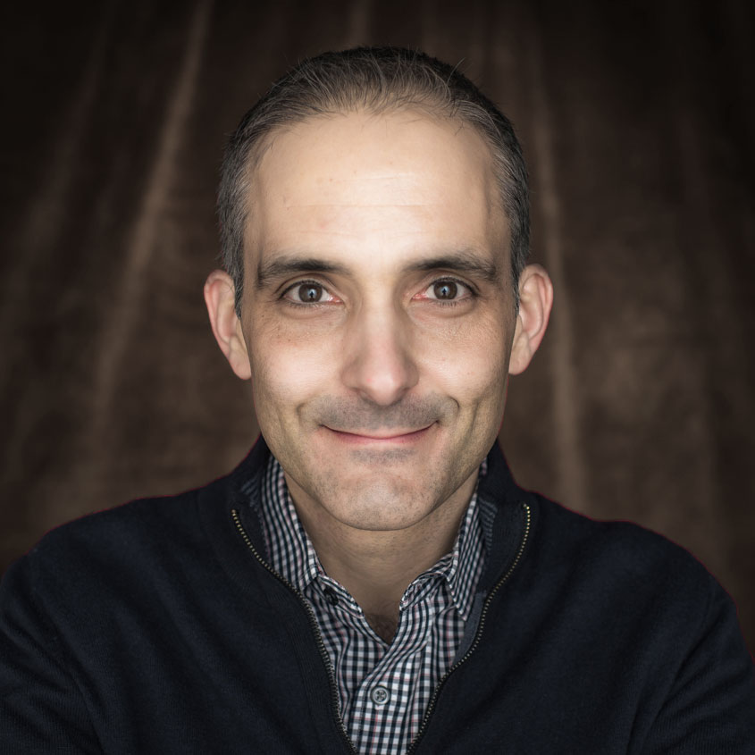 A man with black and grey hair, a medium/olive complexion and wearing a sweater jacket over a checkered, button-down dress shirt stairs ahead at the viewer and smiles