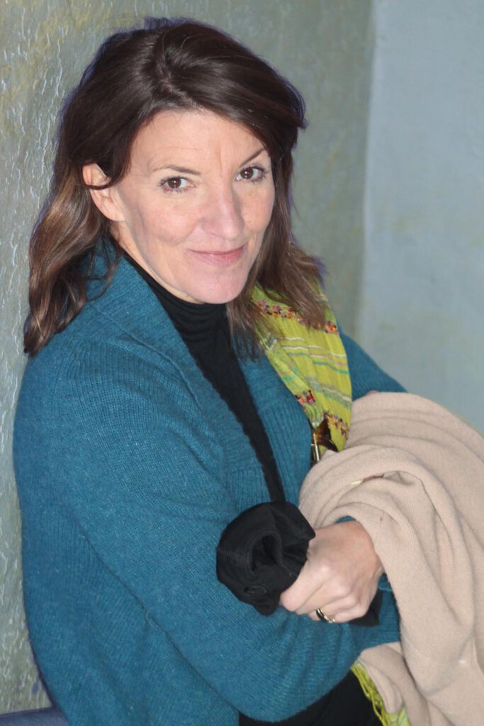 A woman with dark, shoulder length hair parted toward the side, with a fair complexion and wearing a blue sweater jacket and light-green scarf with a pattern