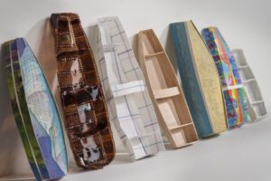 A row of miniature rowboats leaning against a wall, each made of different materials – photo negatives, watercolor paintings, shirt material, thin wood, a written page