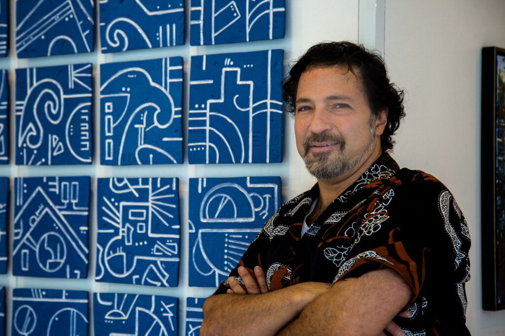 A man with black, wavy hair and a salt-and-pepper goatee with a medium complexion is seen wearing a black short-sleeved shirt with a white and brown floral pattern and is standing in front of a series of blue and white tiles