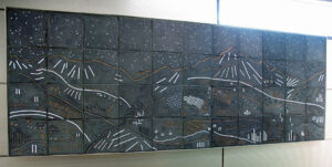 A grid of dark-slate ceramic tiles which, seen together, depict a mountain and landscape scene through stylized white and orange carved lines