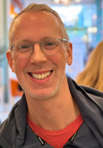 A man with short, light-grey and light-brown hair parted to the side with a slight, white stubble and a fair complexion wearing transparent-framed glasses, a red shirt and windbreaker is seen smiling