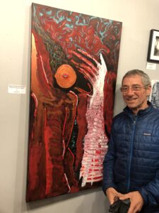 A man with short chair, thin wireframe glasses and a blue winter jacket stands next to a red painting with a hand-like shape and orange shape