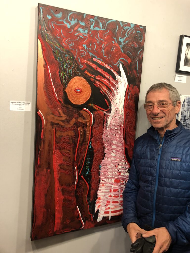 A man with short chair, thin wireframe glasses and a blue winter jacket stands next to a red painting with a hand-like shape and orange shape