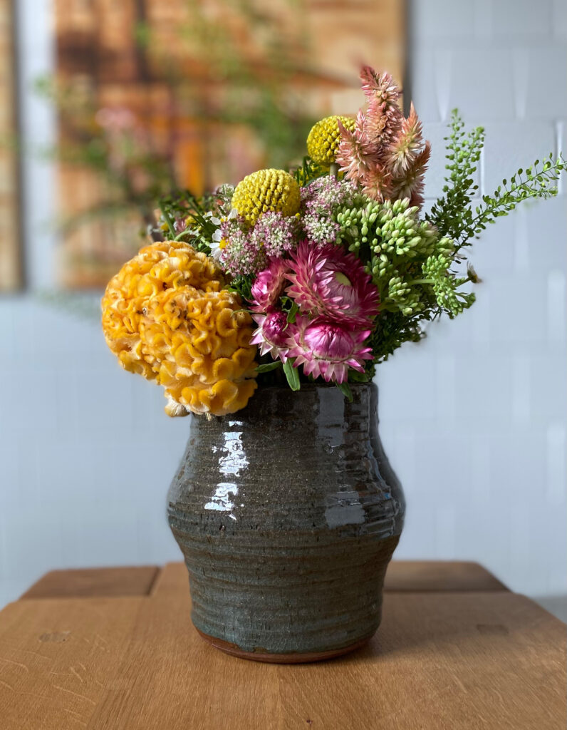 A ceramic, dark olive-green vase with a shiny, banded surface holds an assortment of colorful flowers