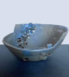 A ceramic bowl with an irregular, shiny surface and shorter on side than the other is seen with a number of small ceramic petals adorning its side