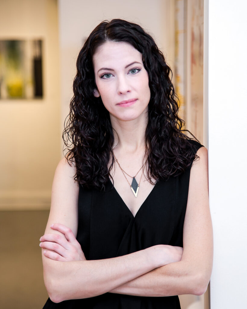 A woman with dark, wavy shoulder-length hair and wearing a black dress and a diamond shaped pendant necklace is seen in an art gallery with arms crossed looking straight ahead toward the camera