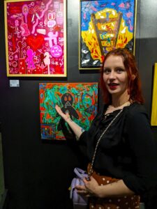 A woman with red, shoulder length hair and wearing black blouse and brown skirt is standing in front of several small paintings on display on a black wall in an art gallery setting.