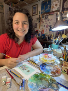 A woman with dark, shoulder-length, wavy-curly hair, light-tan complexion and wearing a red t-shirt is working at a studio table on watercolor paintings and is surrounded in a room by various painting tools and materials