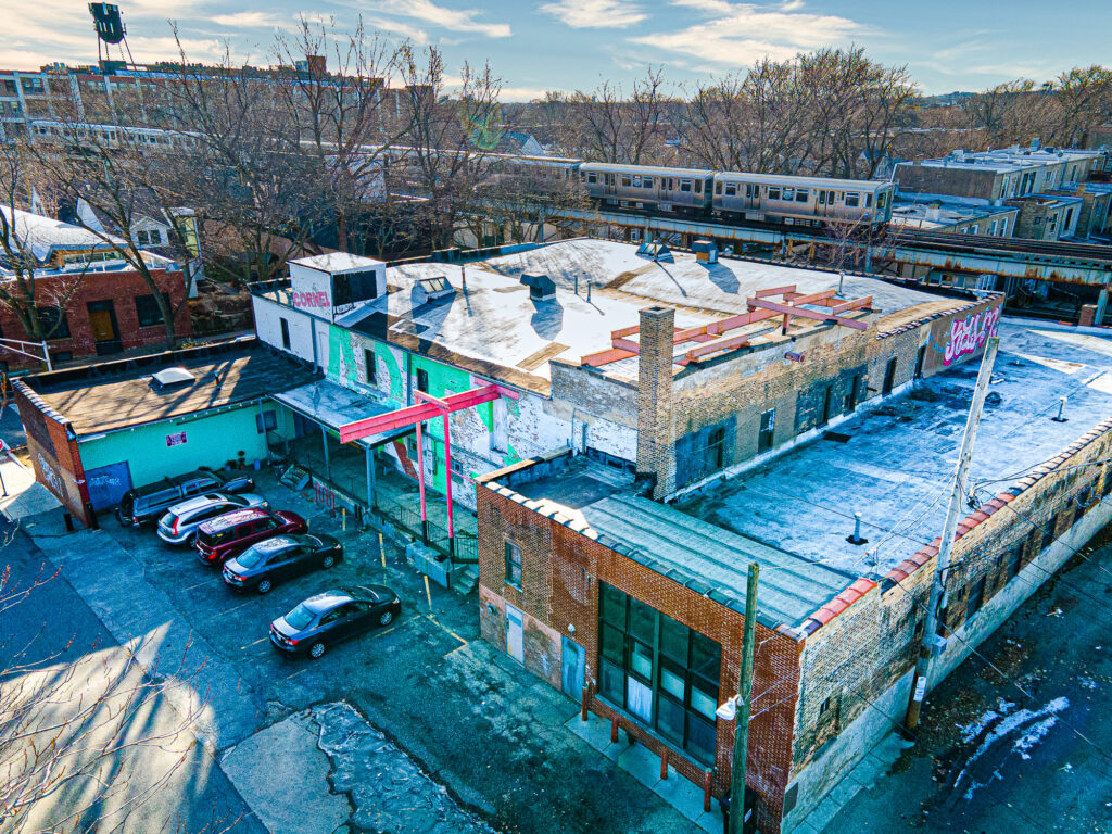An overhead aerial photo of an industrial-looking building in the middle of an urban area. Just behind the building, an elevated public train can be seen moving behind it. The roof of the building has steel beams and skylights covering it