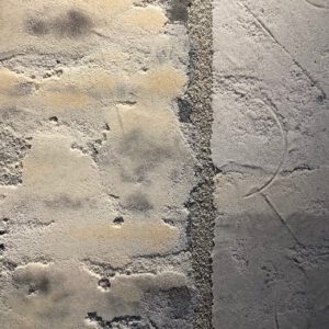 A close-up of a light-gray abstract piece made from various building and earth materials applied in different sections in more or less geometric lines and patterns. The textures are both concrete and sand-like