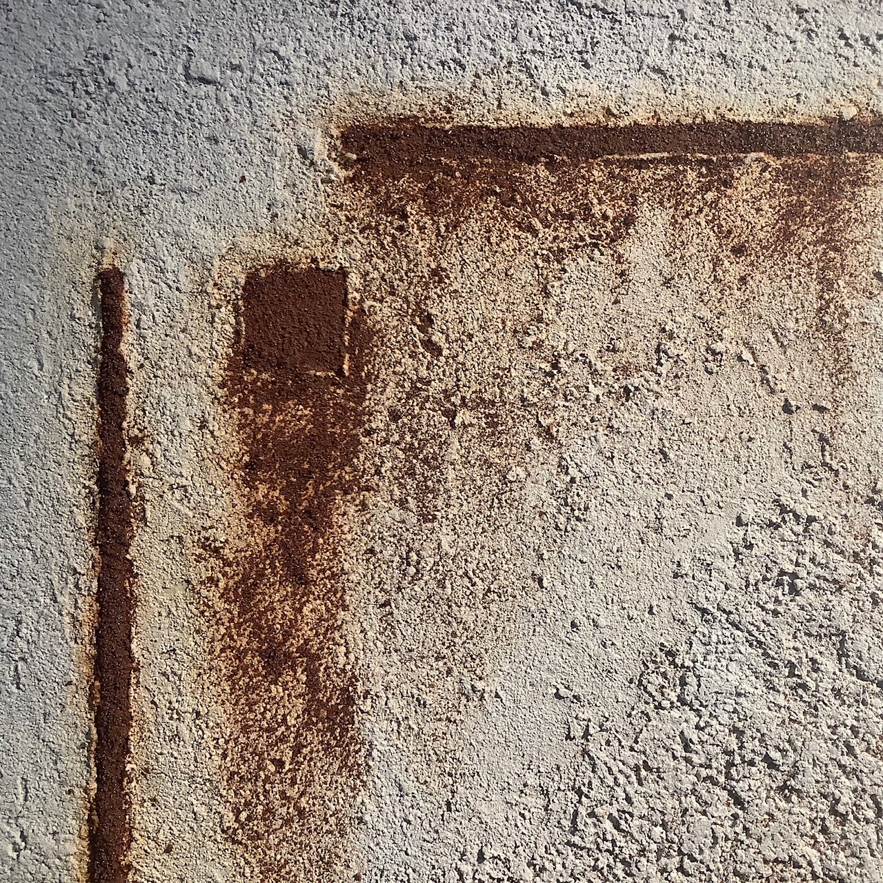 A close-up of an abstract artwork made from building materials. The texture is a beige and gray concrete-like substance with streaks of darker rust-colored powder-like paint material forming rough squares and lines at right angles.