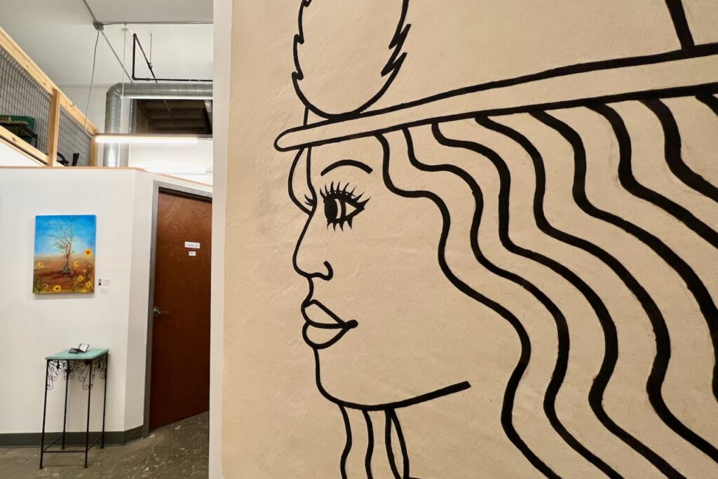 A large, very stylized line drawing of a woman's head is seen in profile on most of the right side of the photo. On the left side, seen further back are the door and wall to an art studio. A small, colorful painting of a tree in a field is hung on the wall with a small end table placed just below