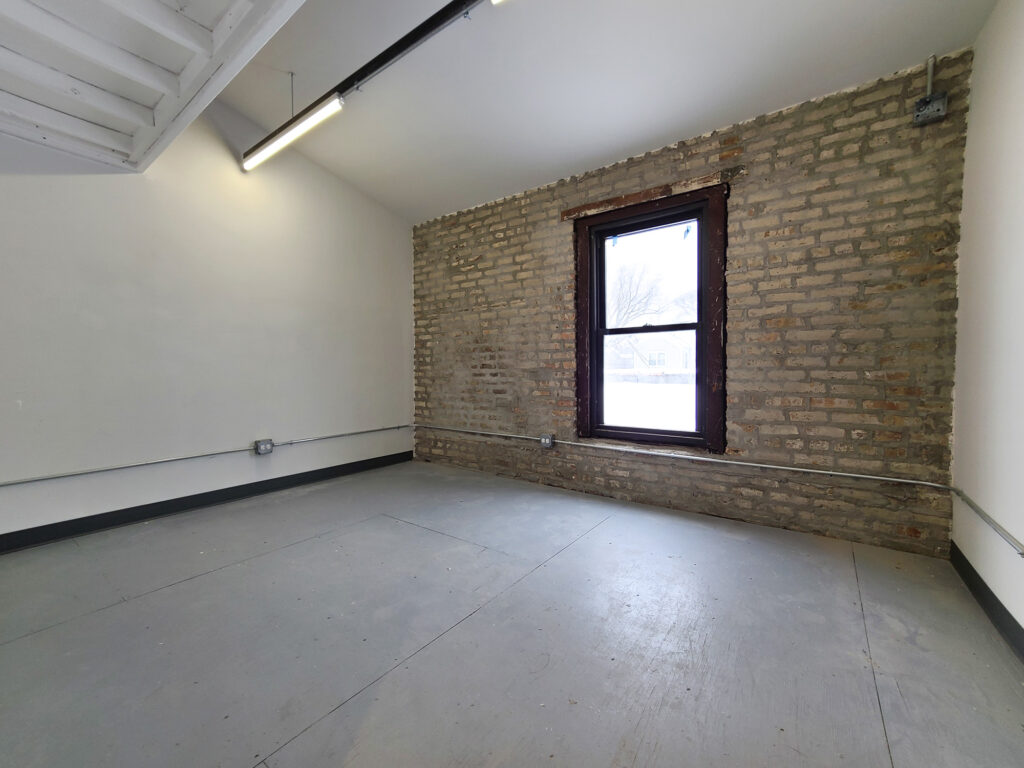 A lofted space with white, freshly-painted studio walls adjoining a brick exterior wall with a large inset window