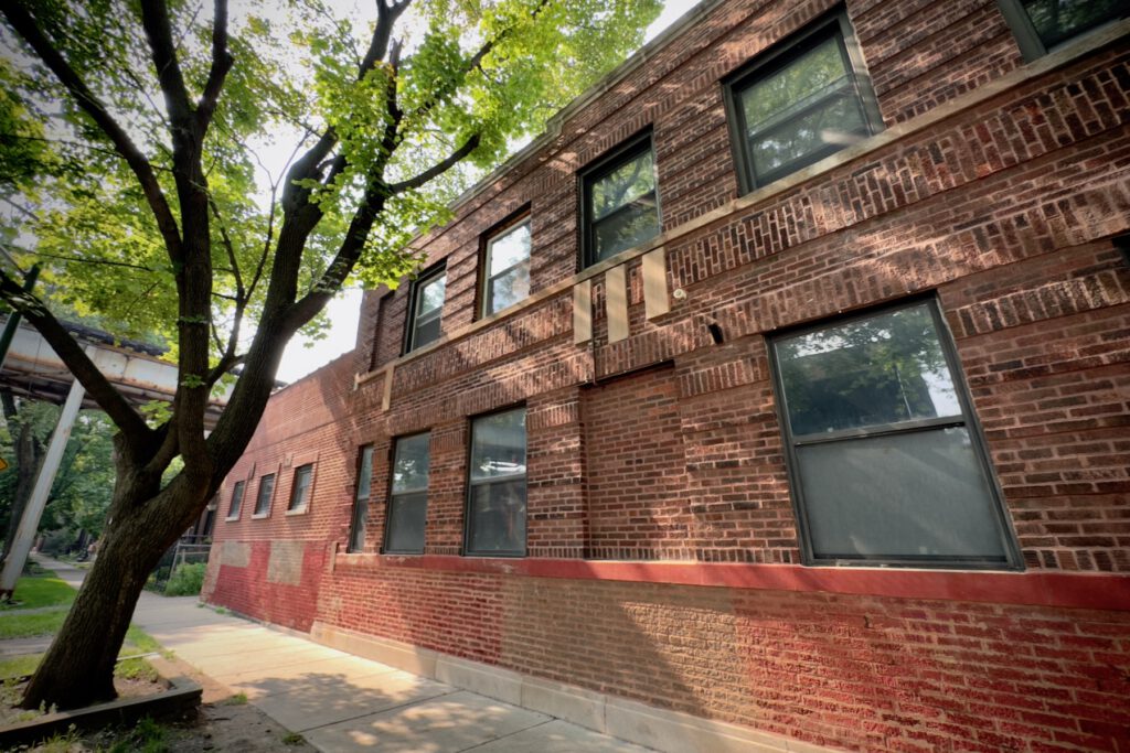 A brown, 2-story brick building on a sunny day along a city street. Elevated train tracks are visible just behind it.