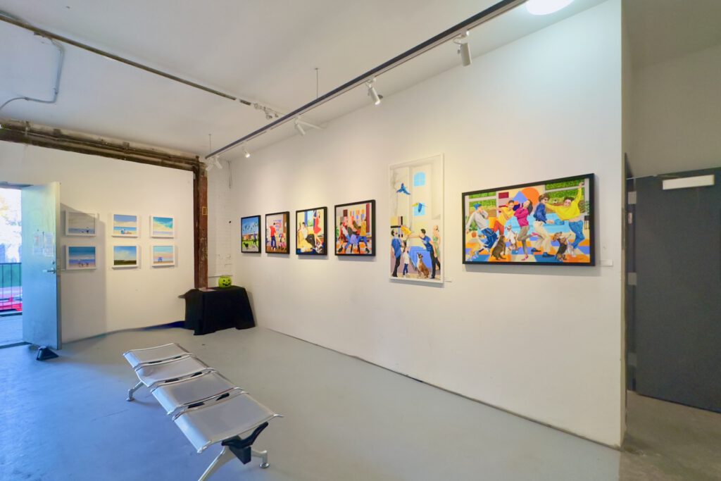 A large, brightly-lit room with white walls and several colorful paintings hung on two separate walls that face the viewer. A stylish metal bunch is placed in the middle of the viewing area
