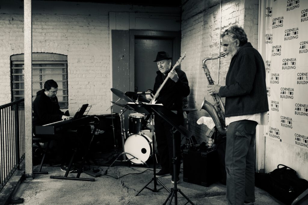 A trio of men play music against the backdrop of a brick building. One plays a keyboard, the other a bass guitar and in the foreground a man plays a saxaphone. Just barely visible behind the trio is a man playing a drum kit