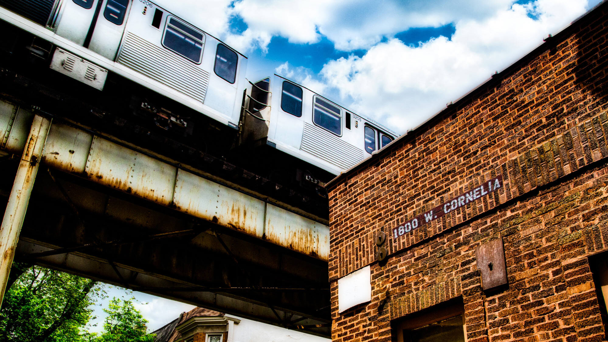 Seen from below, a public train passes by a brick building on elevated train tracks on a sunny day with some clouds and green trees visible in the background