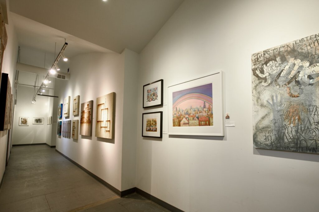 A long, brightly-lit art gallery hallway is shown with an array of pieces hung along the wall by several different artists.