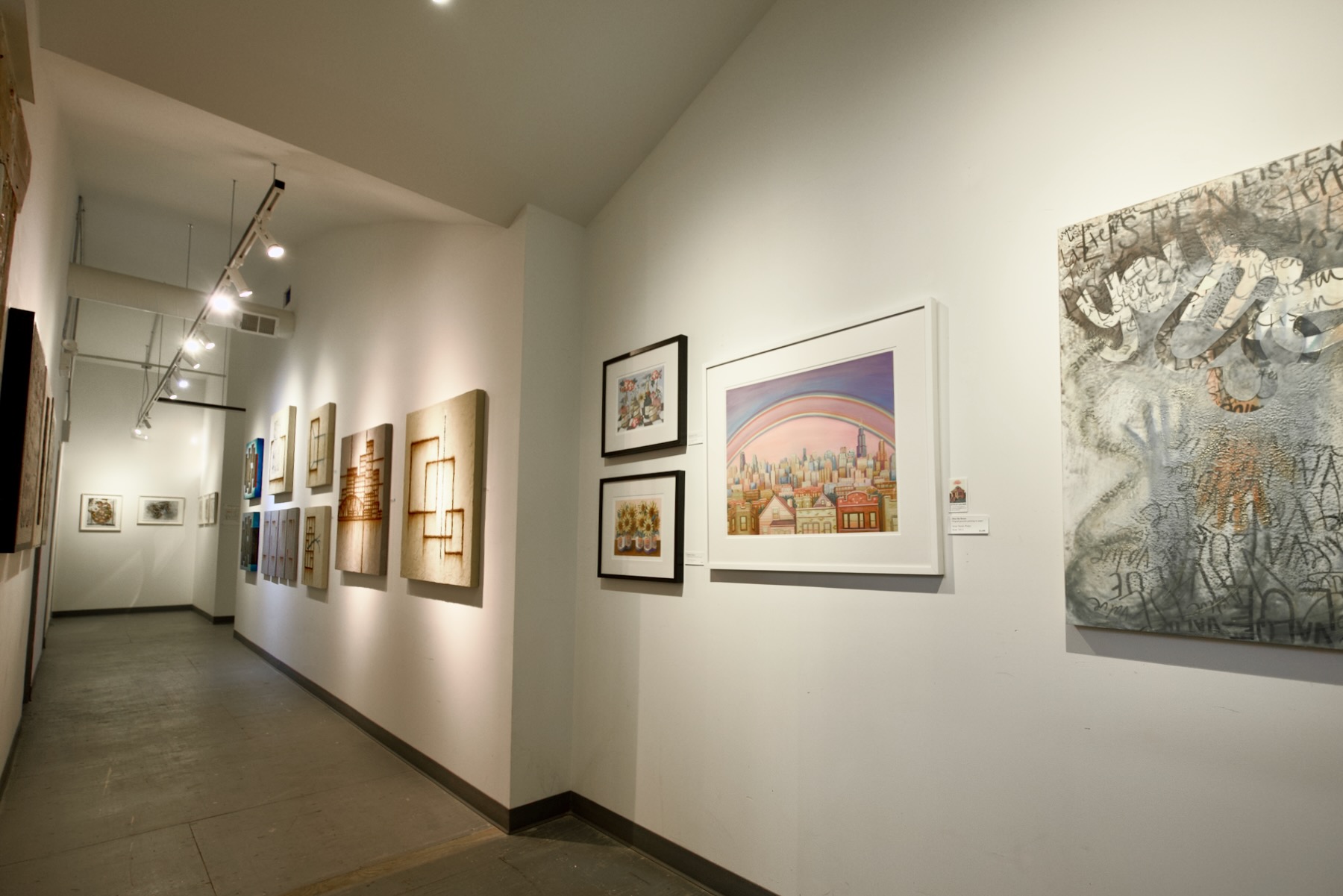 A long, brightly-lit art gallery hallway is shown with an array of pieces hung along the wall by several different artists.