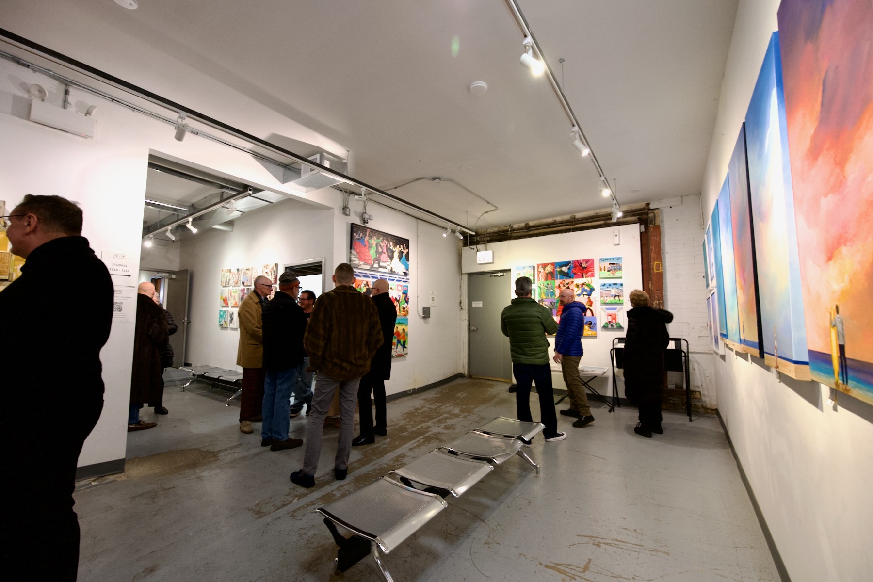 A large, brightly-lit room with white walls and several colorful paintings hung on two separate walls that face the viewer. A stylish metal bunch is placed in the middle of the viewing area. Several people are seen mingling and viewing the artwork