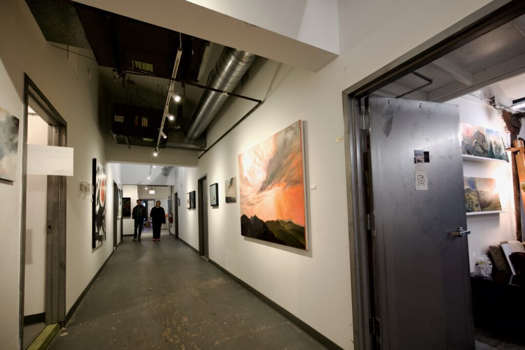 A long hallway in a gallery setting shows an array of paintings hung in between doorways to studios. To the very right, an open door reveals some landscape art and painting supplies just inside a studio.