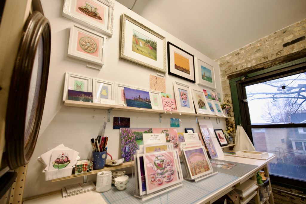 An art studio shows an assortment of paintings of different subject matter including Chicago cityscapes, tea sets, desserts and flowers. Just outside, a city street and house are visible below.
