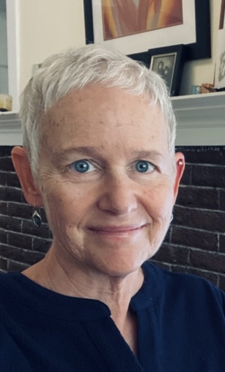A woman with short, grey hair, blue eyes and a fair complexion wearing earrings and a dark blue shirt