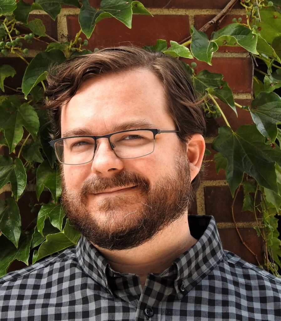 A man with a fair complexion, brown, wavy hair and a medium beard wearing glasses and a grey and black checked shirt stands against a brick wall with ivy growing on it