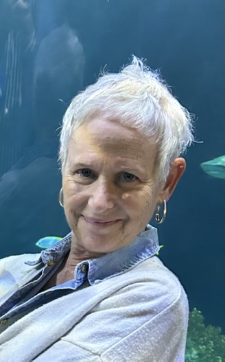 A woman with short, white hair and fair complexion wearing hoop earrings and a light blue denim shirt looks toward the camera