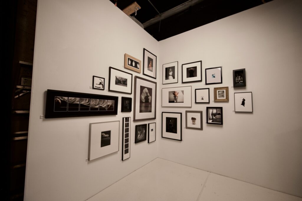 A group of framed photographs of various sizes are hung on a wall corner facing toward one another in a stark white gallery wall set a against a dark room background
