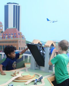 A museum exhibit for children is a city-like setting with a wooden drawbridge and toy train track pieces on a table. Two young boys play with the drawbridge