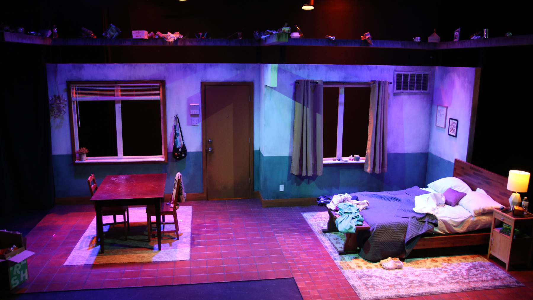 A stage production set of a worn down, small studio apartment that has a simple table and 2 chairs as furniture and a messy bed is illumintated with blue and purple stage lighting