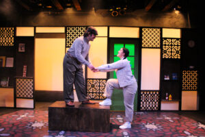A male actor in a hat, dress pants, and long-sleeved grey turtleneck stands on a podium bending down to a female actor with a sweater, long brunette hair and dress slacks on. They are on a stage with production lighting and a living room-like set shown behind them.