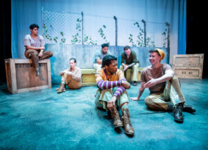A group of actors in a scene on a play production set are dressed in ragged-looking clothes with makeup to make them appear as a group of laborers. A chain link fences and wooden crates are stage props visible throughout the image.