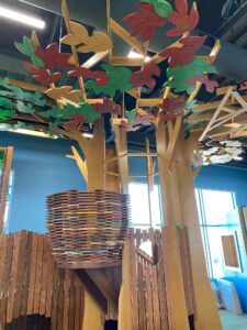 An indoor playground made of wooden slats cut to resemble a tree with red and green leaves
