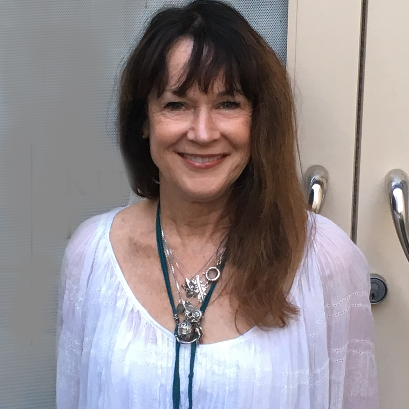 A woman with long brown hair and fair complexion and wearing a white blouse with a group of different necklaces and pendants is smiling toward the viewer