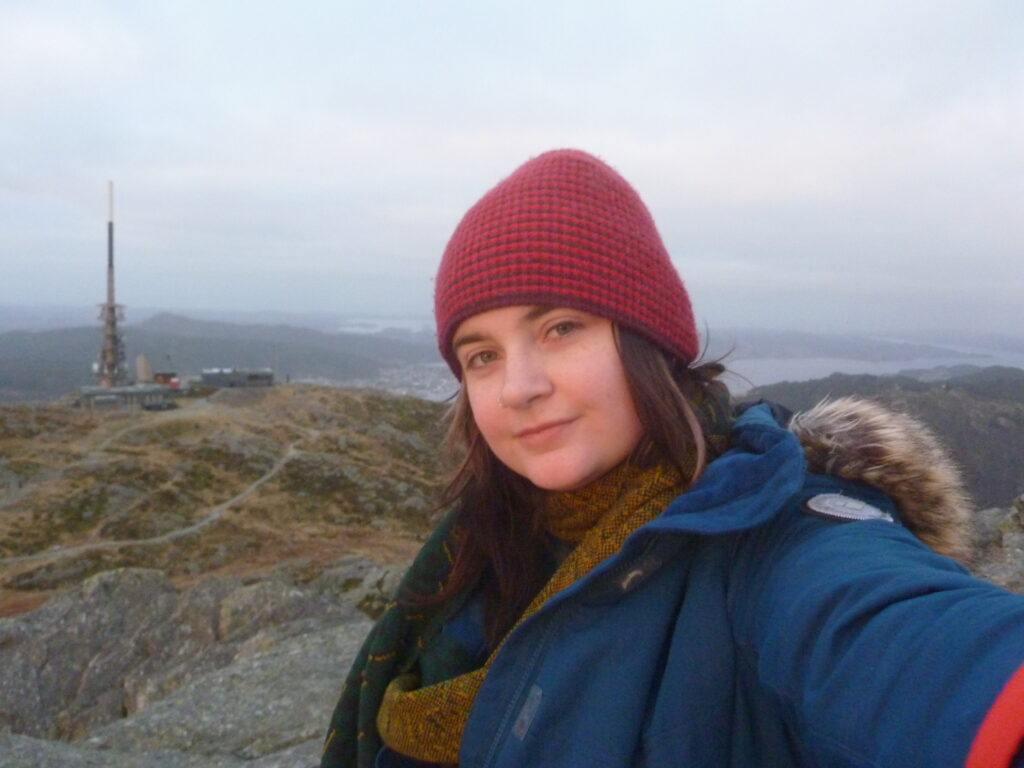 A woman with a fair complexion, brunette hair and wearing a red knitted stocking cap, blue winter coat and patterned scarf is standing on a very hight hilltop with a weather station facility in the background