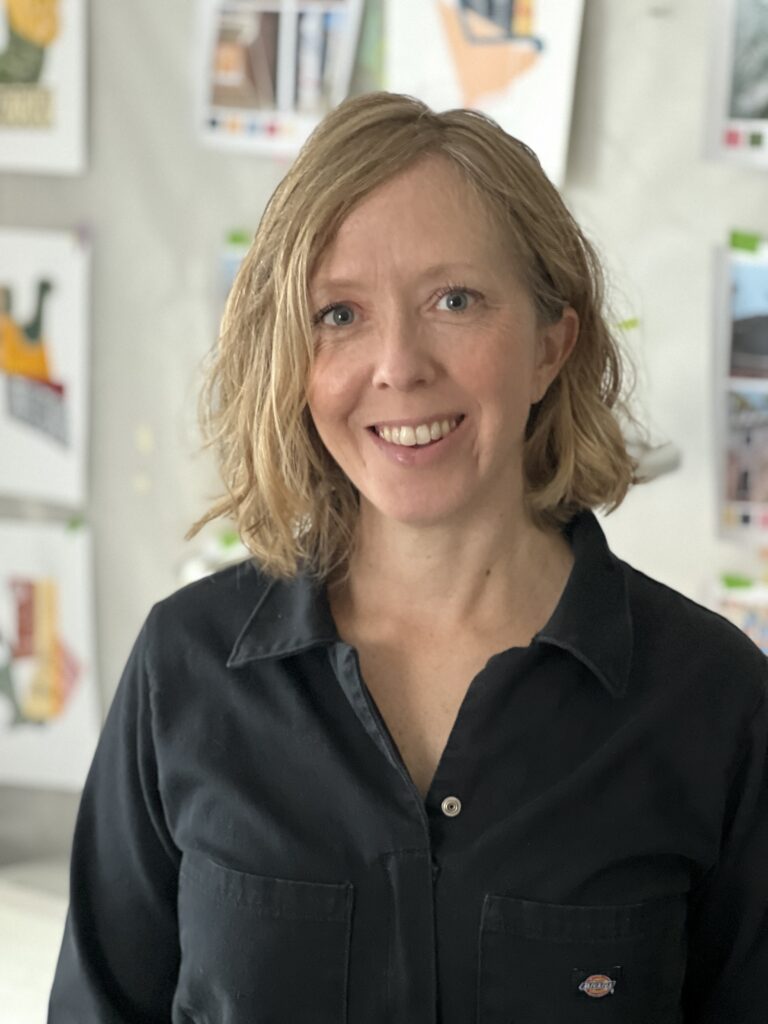 A woman with neck-length, wavy blonde hair wearing a black blouse is standing in a studio with art prints hanging on the wall behind her
