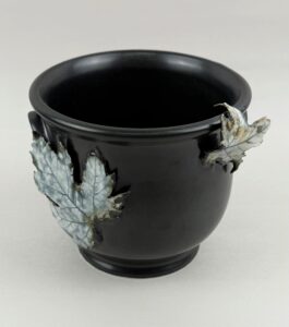 A black ceramic cup with decorative sculptures of leaves with muted green-grey colors attached to the side